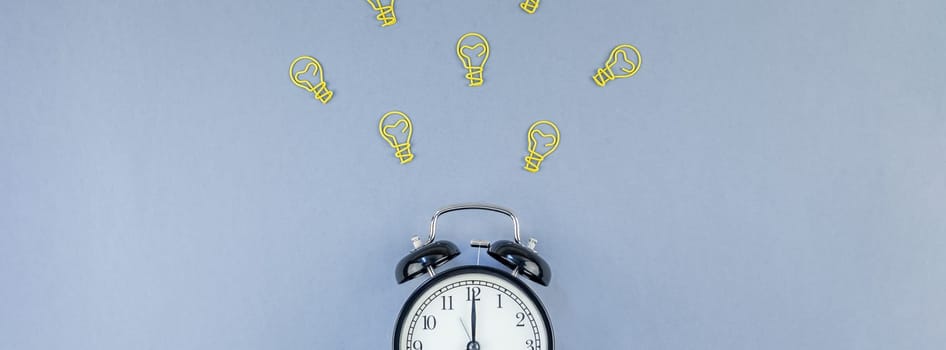 Top view flat lay of desk with black alarm clock and bulbs idea signs with copy space on blue background in minimal style. Concept of creativity, new plans, brainstorming, creative work