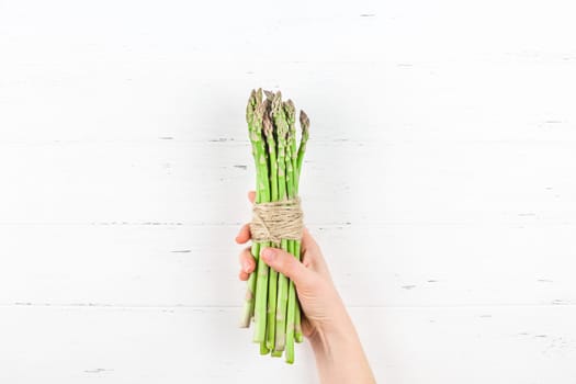 Creative scandinavian style flat lay top view of fresh green asparagus in woman hands on white wooden table background copy space. Minimal house cooking concept for blog or recipe book