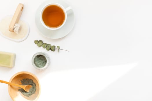 Spa, beauty cosmetics and body care treatment concept with copy space. Creative top view flat lay composition with cup of tea, green spirulina cleansing mask in wooden bowl on white background