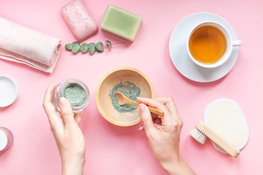 Spa, beauty cosmetics and body care treatment concept copy space. Creative top view flat lay of woman hands with bath accessories, green spirulina cleansing mask in bowl, cup of tea pink background