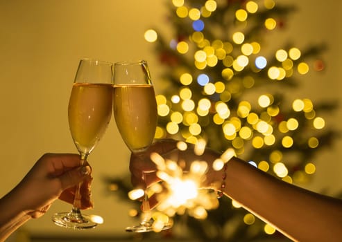 Two hands clinking champagne glasses in celebration party toast with a sparkling bokeh of Christmas lights and festive tree in the background