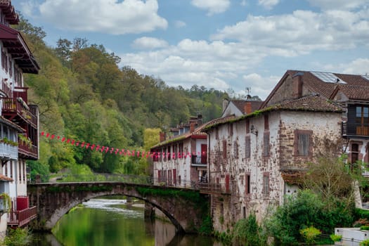 A bridge spans a river in Saint Jean Pied de Port, France. The bridge is decorated with red flags and the town is surrounded by trees