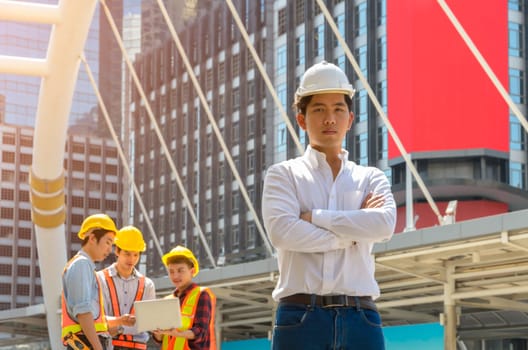 Portrait of Asian male project engineer in white helmet standing with crossed arms in front of modern building with three men working with laptop on background