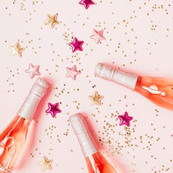 Christmas or New Year composition with bottles of rose champagne and golden shiny sparkle star confetti on pastel pink background, top view. Celebration flat lay. Party creative concept