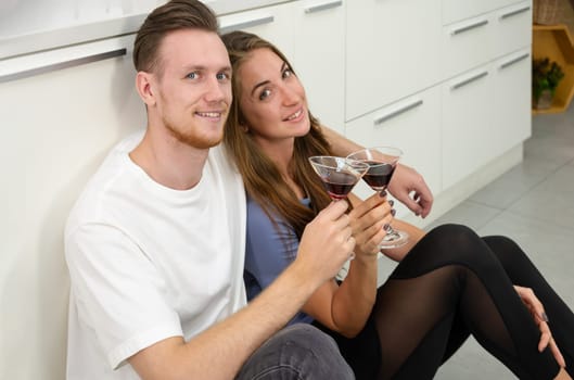 Smiling Caucasian man and woman sitting on kitchen floor and holding glasses of cocktails. Romantic couple spending time together, having fun and enjoy drinking at home.