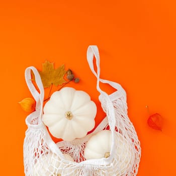 Autumn flat lay composition with white pumpkins in mesh shopping bag on bold orange color background. Creative autumn, thanksgiving, fall, halloween concept in zero waste style. Top view, copy space