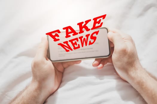 Fake news infodemics during Covid-19 pandemic concept. Man hands holding mobile phone reading global news or social net at home. Reading last news update in lockdown. Red text fake news on foreground
