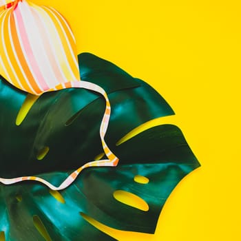 Tropical jungle palm monstera leaves and a woman's swimsuit top on bright yellow square paper background. Summer vacations creative flat lay concept template for text