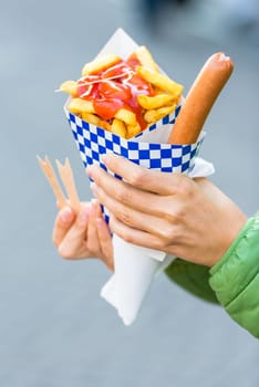 Female hands holding paper cone with a hot sausage and french fries with ketchup