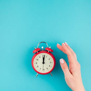 Creative flat lay concept top view of woman hand holding the red vintage alarm clock on square bright blue turquoise color paper background with copy space in minimal style, template for text