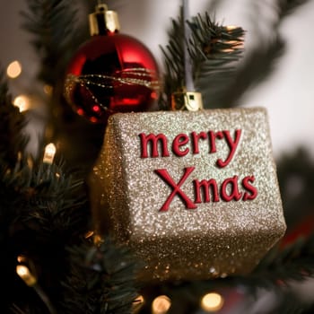 A glittery gold gift tag with Merry Xmas text hangs on a decorated evergreen tree with red ornaments and twinkling lights.