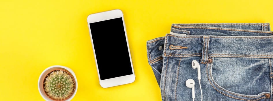 Flat lay of blue jeans, cactus and smartphone headphones on bold yellow paper background with copy space. Overhead view of woman casual outfit. Long wide banner