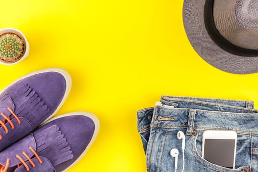 Flat lay of suede shoes, blue jeans, grey hat, cactus and smartphone headphones on bold yellow paper background with copy space. Overhead view of woman casual outfit. Trendy hipster look top view