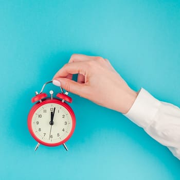 Creative flat lay concept top view of woman hand in white shirt holding the red vintage alarm clock on bright blue turquoise square color paper background with copy space in minimal style, template