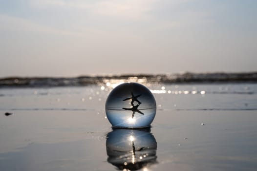 A glass ball lies in the waves on the sandy beach, the ball reflects the sea, a starfish and the setting sun