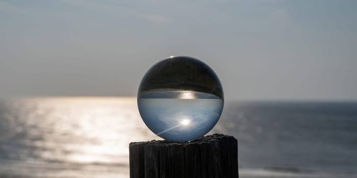 Glass ball lies on a wooden post, in the ball the sea, the sun and the blue sky are reflected