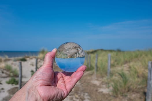 A human hand holds a glass ball, in the ball a sand dune, a footpath, blue sky and the sea are reflected