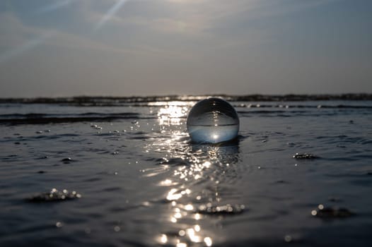 A glass ball lies in the waves on the sandy beach, the sea and the setting sun are reflected in the ball