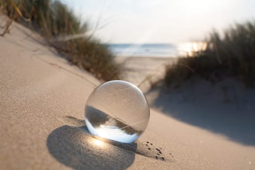 A glass ball lies in the sand , the sea and dune grass  are reflected in the ball