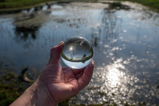 A human hand is holding a glass ball, in the ball a small lake in green nature is reflected, the light of the sun is reflected in the water