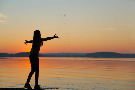 A young girl joyfully plays alongside the serene water at sunset, beautifully capturing a moment of pure bliss