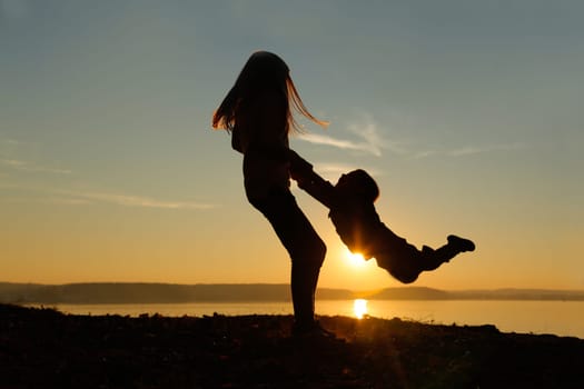The silhouette perfectly captures the image of a girl lifting her brother in her arms against the background of a sunset by the water