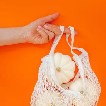 Autumn flat lay composition with white pumpkins in mesh shopping bag on bold orange color background. Creative autumn, thanksgiving, fall, halloween concept in zero waste style. Top view, copy space