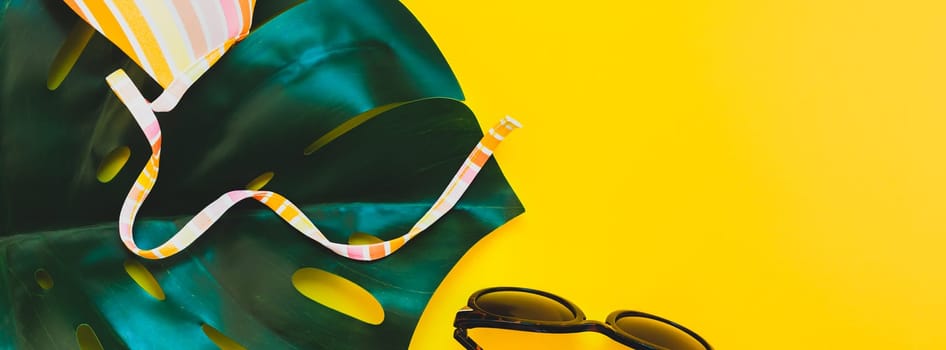 Tropical jungle palm monstera leaves, sunglasses and a woman's swimsuit top on bright yellow paper background. Summer vacations creative flat lay concept template for text. Long wide banner