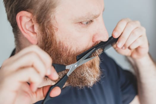 Selfcare during quarantine isolation. Adult handsome caucasian man cutting moustache and beard himself personally with scissors at home. Coronavirus outbreak and people shutdown, stay home concept