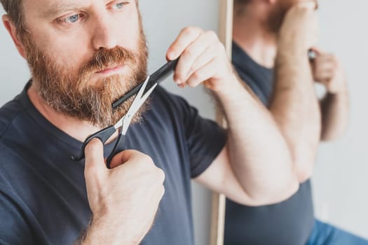 Selfcare during quarantine isolation. Adult handsome caucasian man cutting moustache and beard himself personally with scissors at home. Coronavirus outbreak and people shutdown, stay home concept