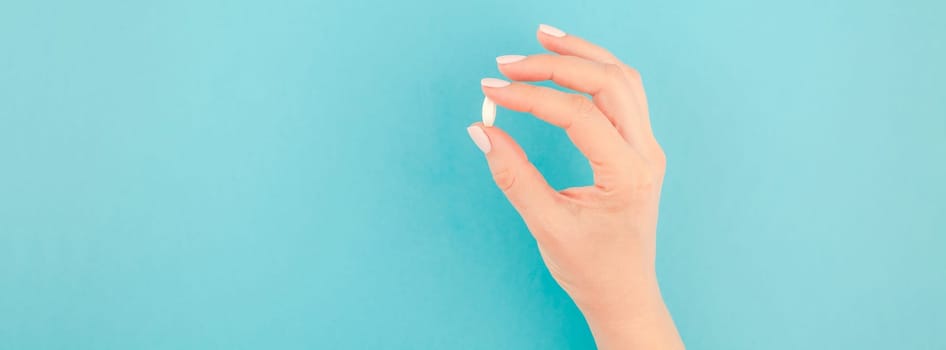Woman hand holding a white pill on pastel blue paper background with copy space in minimal style, template for text. Concept of medical treatment, pharmacy, healthcare. Long wide banner