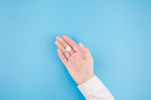 Woman hand holding a white pill on pastel blue paper background with copy space in minimal style, template for text. Concept of medical treatment, pharmacy, healthcare.
