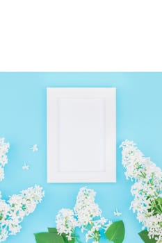 Creative flat lay concept top view of blank postcard frame mock up and white lilac flowers petals on pastel blue background with copy space in minimal style, template for lettering, text or design