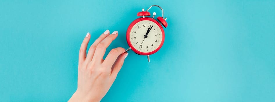Creative flat lay concept top view of woman hand holding the red vintage alarm clock on bright blue turquoise color paper background with copy space in minimal style, long wide banner