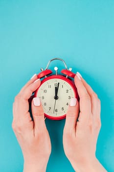 Creative flat lay concept top view of woman hands holding the red vintage alarm clock on bright blue turquoise color paper background with copy space in minimal style, template for text