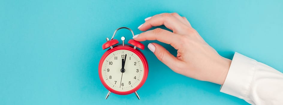 Creative flat lay concept top view of woman hand in white shirt holding the red vintage alarm clock on bright blue turquoise color paper background with copy space in minimal style, long wide banner