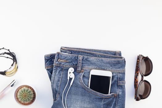 Flat lay of suede shoes, blue jeans, bracelets and smartphone with headphones on white background with copy space. Overhead view of woman casual outfit. Trendy hipster look top view