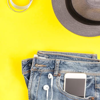 Flat lay of suede shoes, blue jeans, grey hat, cactus and smartphone headphones on bold yellow paper background with copy space. Overhead view of woman casual outfit. Square hipster look top view
