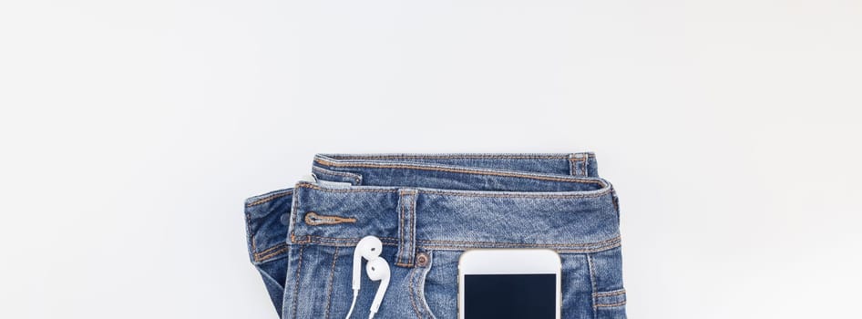 Long wide banner Flat lay of blue jeans and smartphone headphones isolated on white background with copy space. Overhead view of woman casual outfit. Trendy hipster look top view