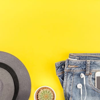 Flat lay of blue jeans, grey hat, cactus and smartphone headphones on bold yellow paper background with copy space. Overhead view of woman casual outfit. Square hipster look top view