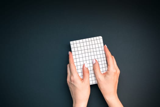 Top view of woman hands with stylish manicure holding notepad with copy space on black color paper background in minimal style. Template for feminine blog social media