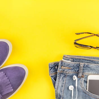 Flat lay of suede shoes, blue jeans, grey hat, cactus and smartphone headphones on bold yellow paper background with copy space. Overhead view of woman casual outfit. Square hipster look top view