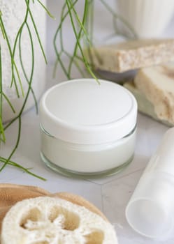 White cream jar with blank lid on white marble against potted green plant and soap bars in bathroom, close up, packaging mockup. Natural cosmetic product for facecare, beauty routine