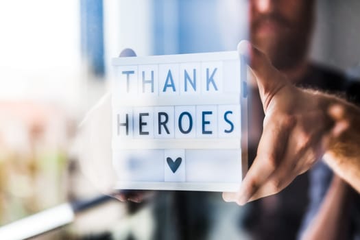 Nurse day concept. Man hands holding lightbox with Thank heroes text thanking doctors, nurses and medical staff working in hospitals during coronavirus COVID-19 pandemics. View through window glass
