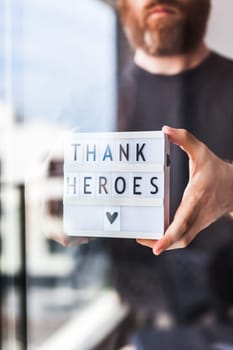 Nurse day concept. Man hands holding lightbox with Thank heroes text thanking doctors, nurses and medical staff working in hospitals during coronavirus COVID-19 pandemics. View through window glass