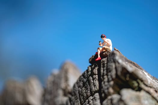 Miniature man sitting on a rock using a smartphone and listening music in headphones with a blue sky background
