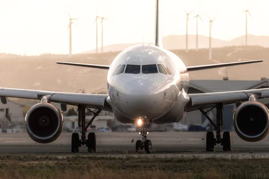 Modern passenger airplane is on the runway at sunset with wind turbines in the background