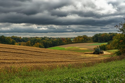 Panoramic view of diverse farmland with harvested and green fields, tractor at work, under dramatic cloudy sky, framed by autumn trees.