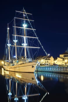 Sailing boat anchored in old town of Klaipeda, Lithuania, with evening lights on. Medieval town and barquentine reflection in calm waters.