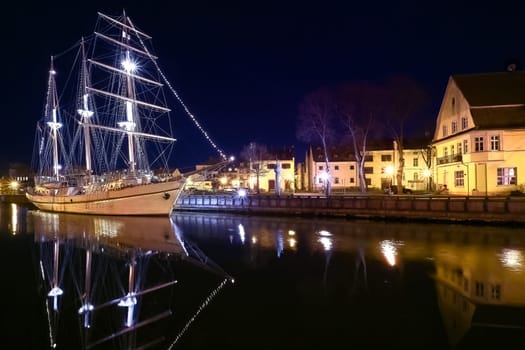 Sailing boat anchored in old town of Klaipeda, Lithuania, with evening lights on. Medieval town and barquentine reflection in calm waters.